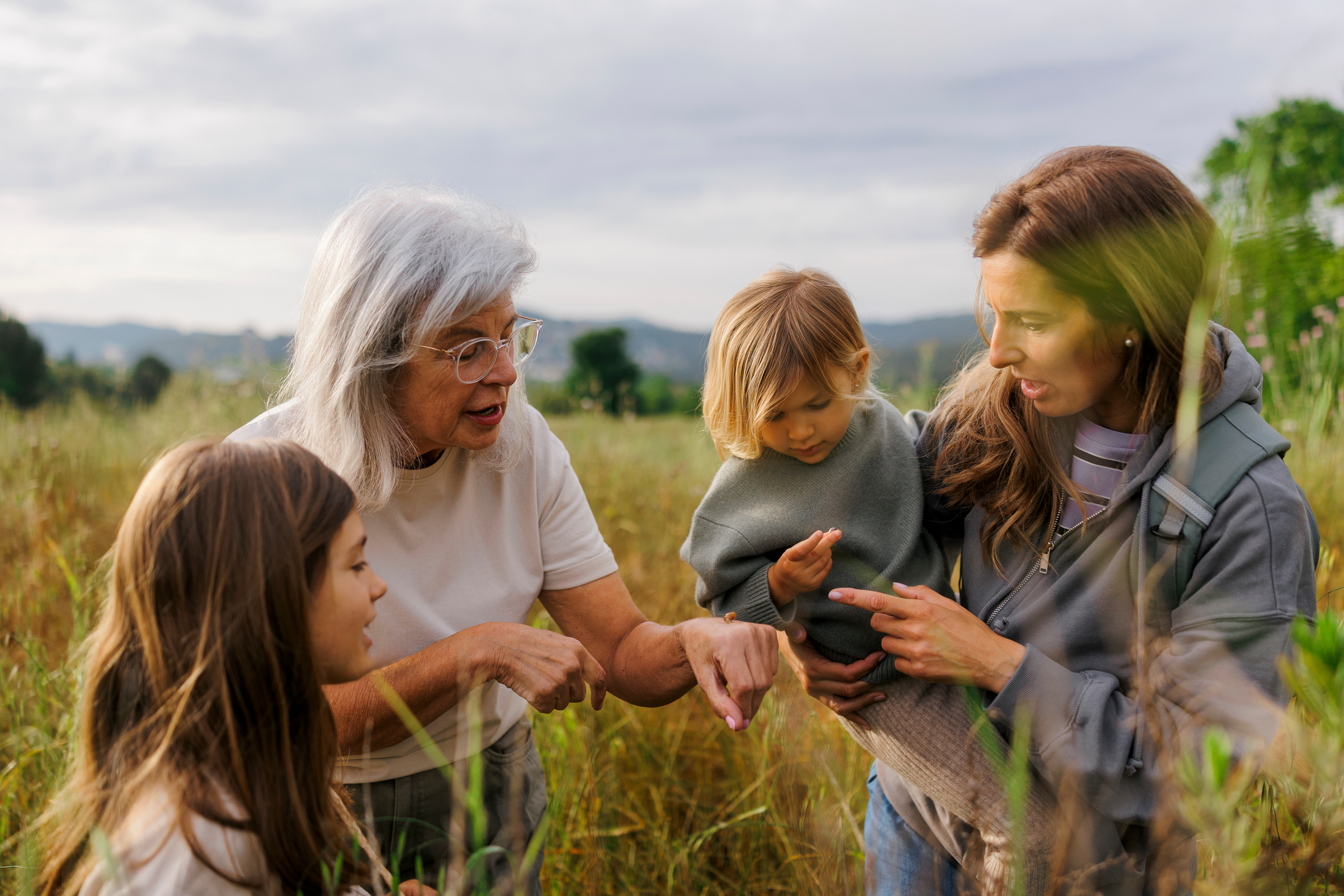 Outdoors in a grassy field, a grandmother, a mother holding a toddler, and a young girl are gathered closely. 