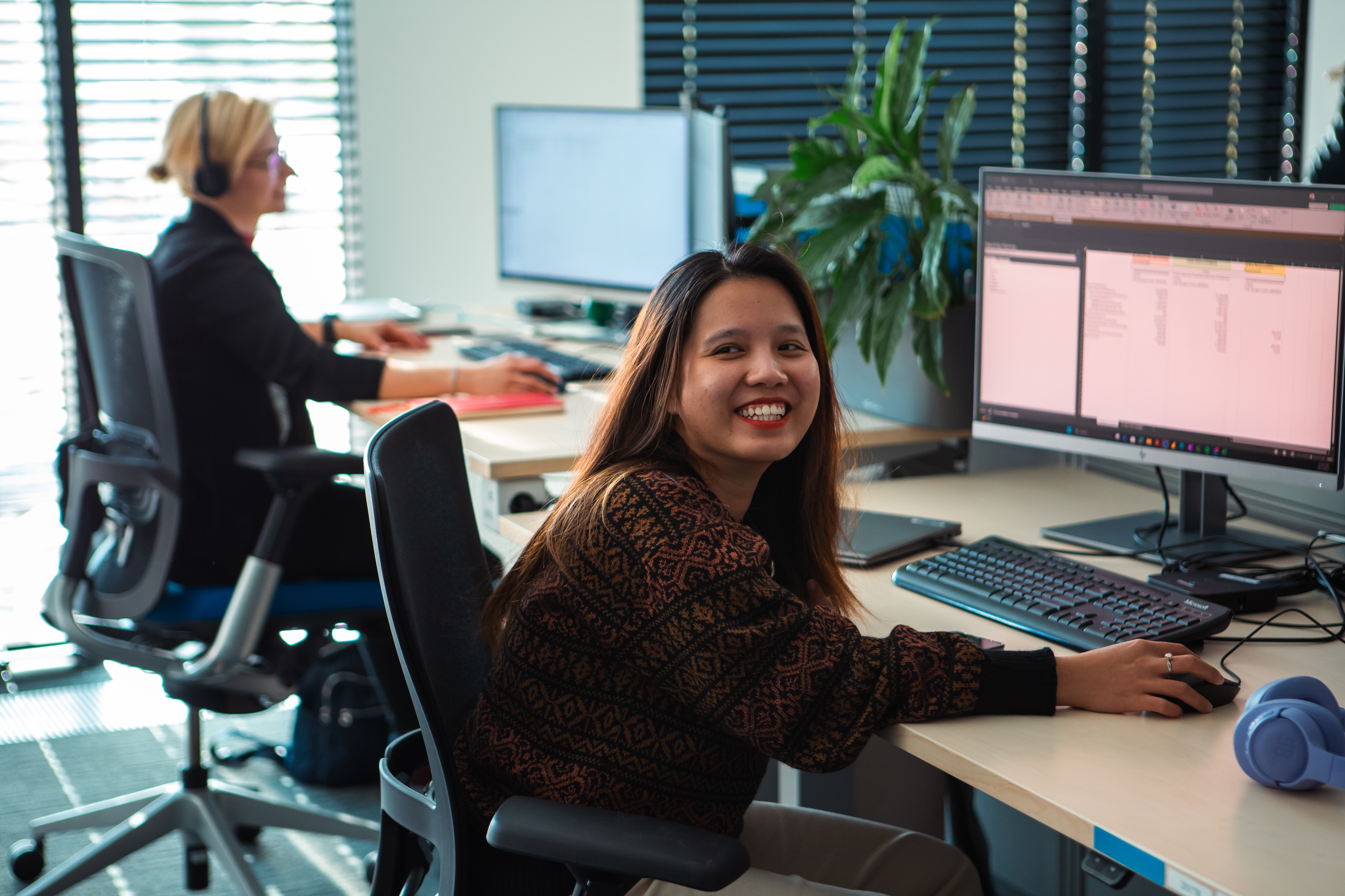 A young woman with long dark hair sits at her desk in a bright, modern office, turning in her chair to smile at the camera. 