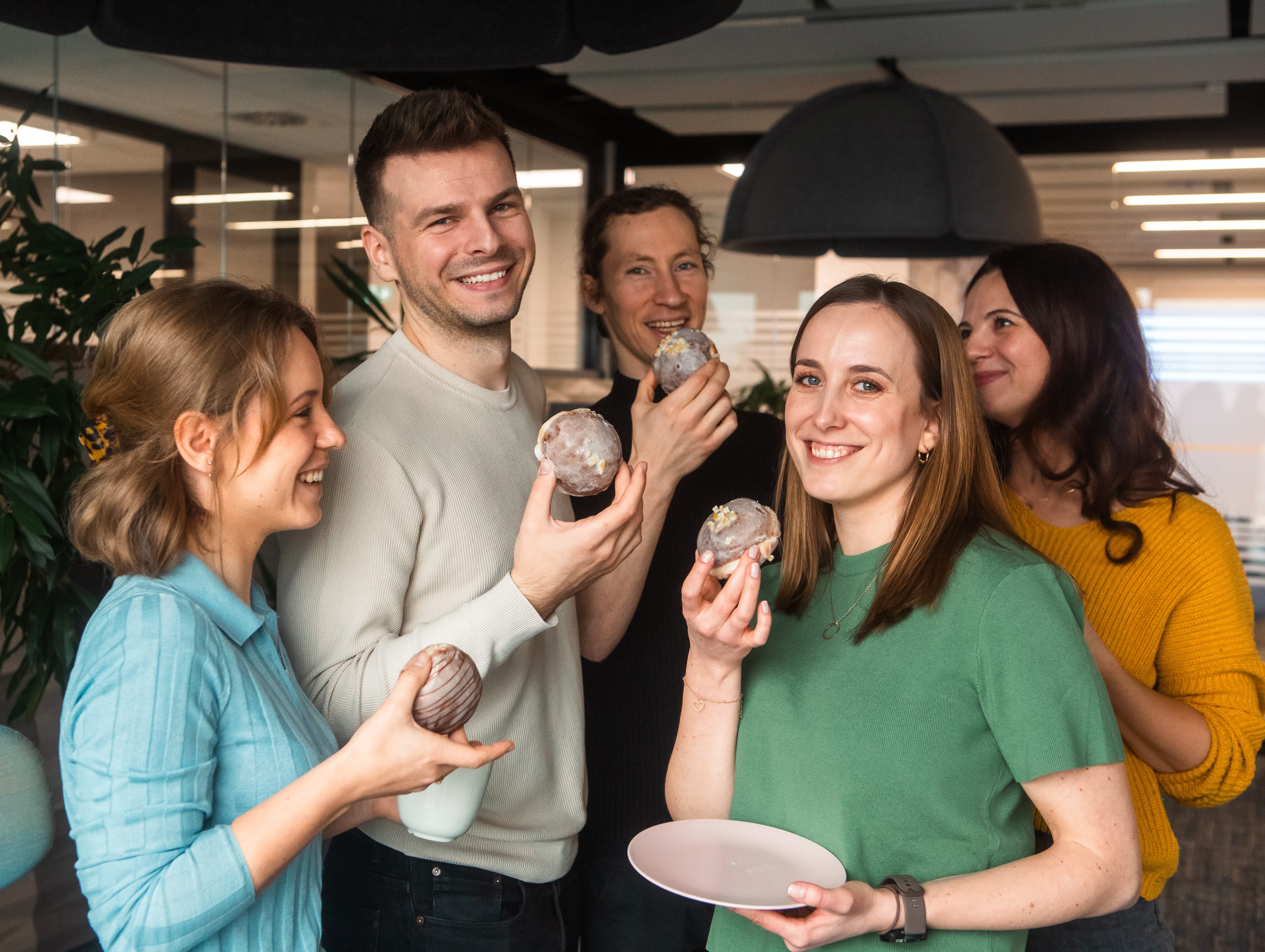 A group of five happy coworkers smiling and eating glazed donuts together in a modern office.
