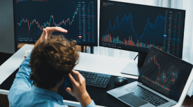 A man in a blue shirt sits at a desk with two large monitors and a laptop, all displaying complex financial charts. 