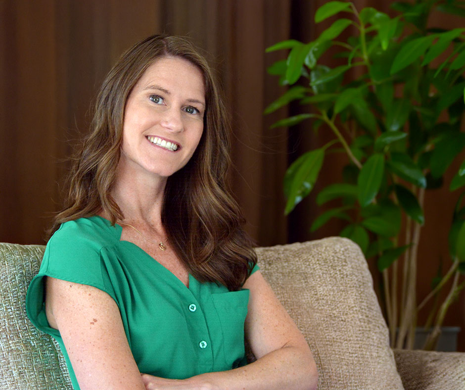 ALT text:A smiling woman with long, wavy brown hair, wearing a vibrant green, short-sleeved blouse, sits on a beige couch with her arms crossed.