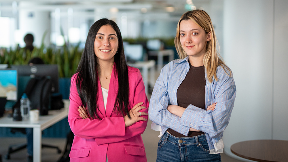 Two individuals stand with arms crossed in a modern office space, surrounded by desks, computers, and greenery.