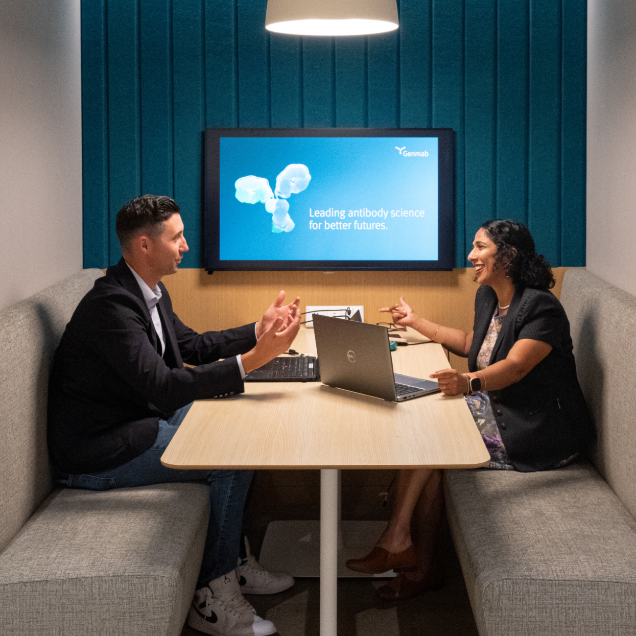 Two people sit across from each other in a modern meeting booth, talking and smiling with laptops open. A screen behind them displays "Leading antibody science for better futures.