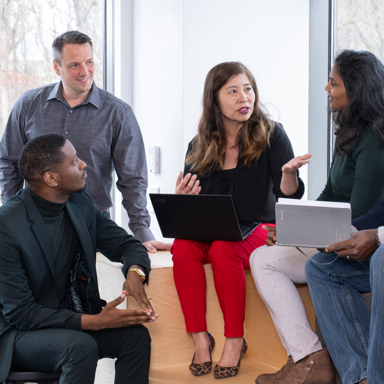Four colleagues sit and stand by a window, engaged in discussion. One woman with a laptop gestures while talking, as the others listen attentively and hold notebooks.