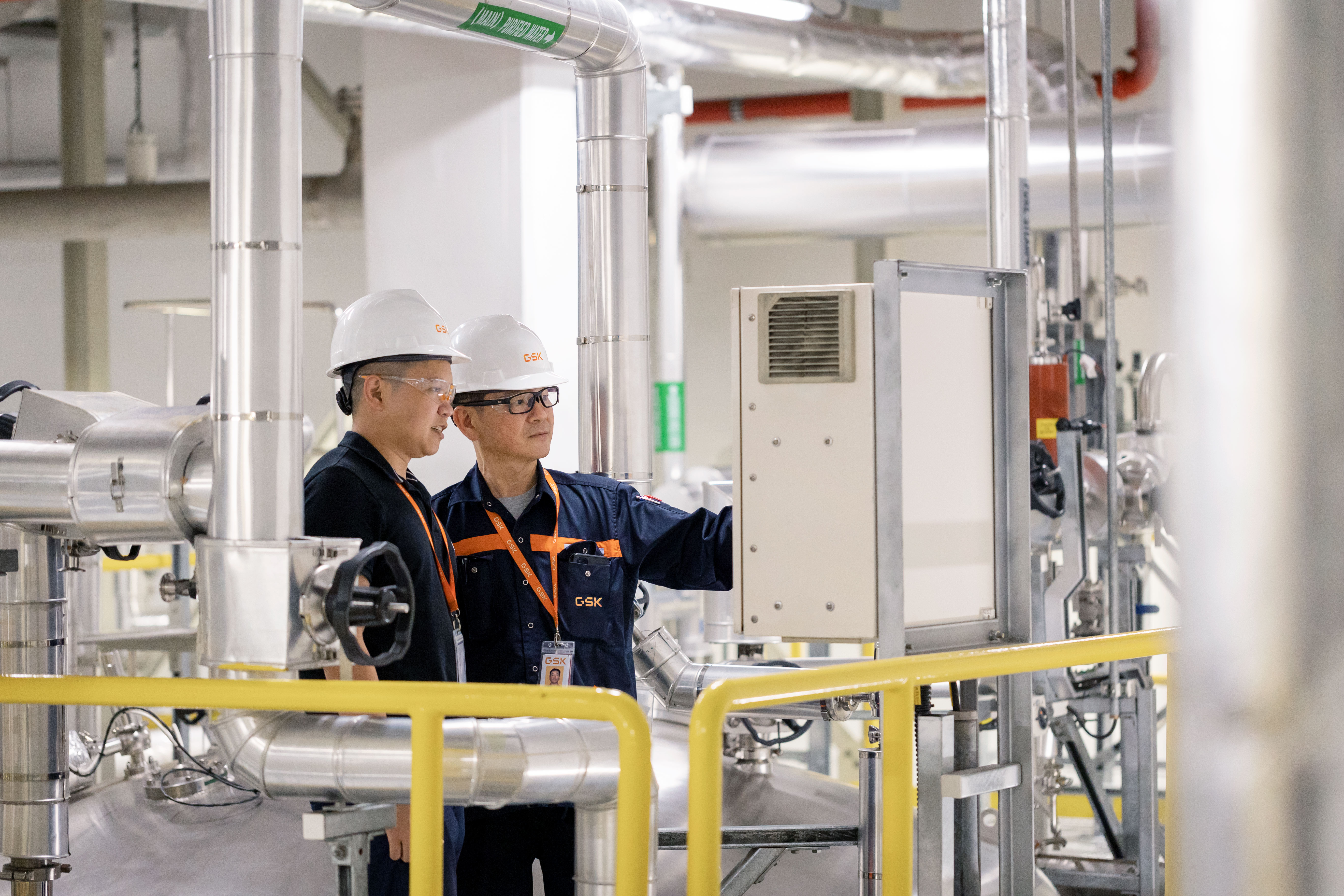 Two workers wearing hard hats and GSK uniforms examine equipment in an industrial facility with pipes and safety railings.