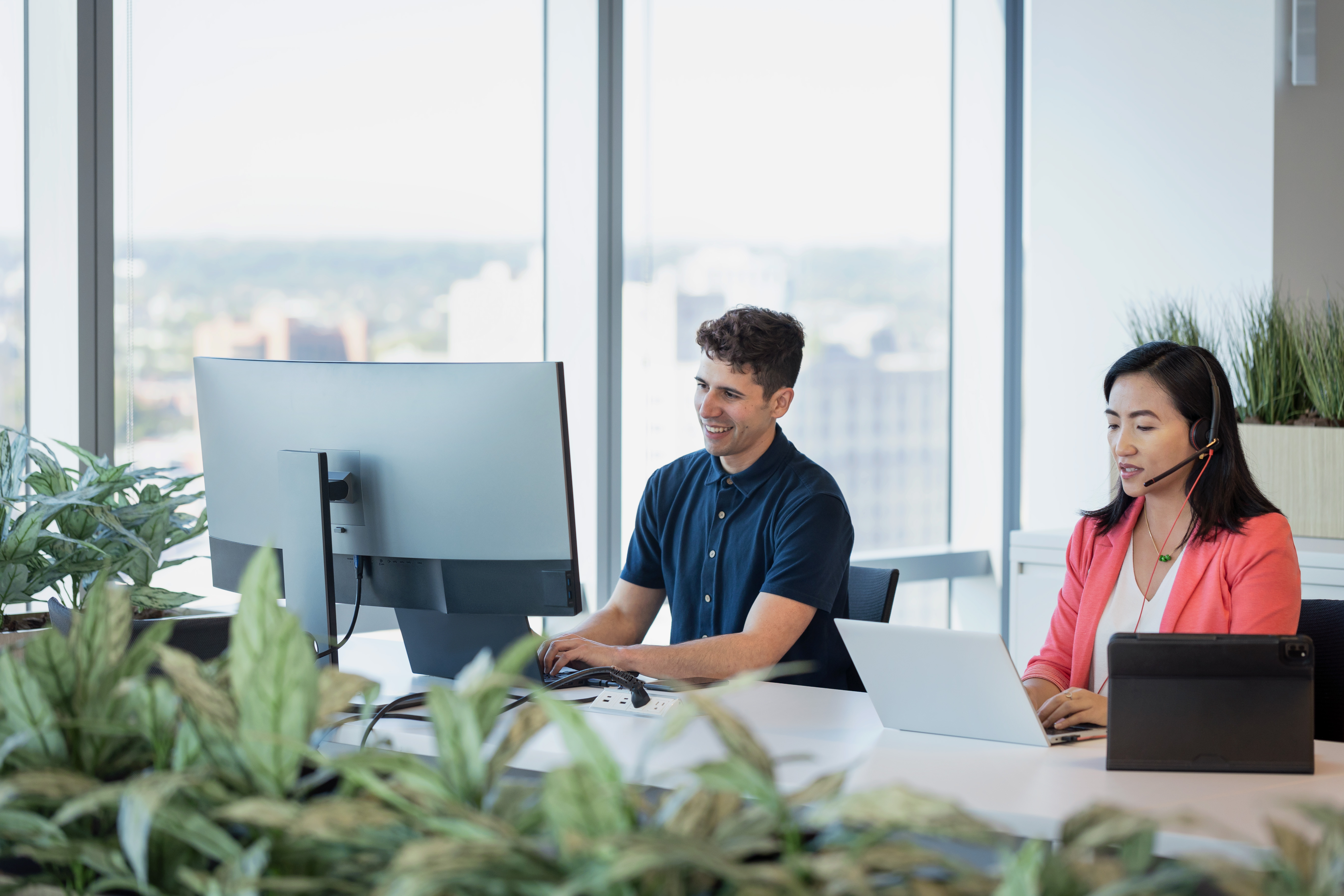 Two people working at computers in a bright office with large windows and plants.