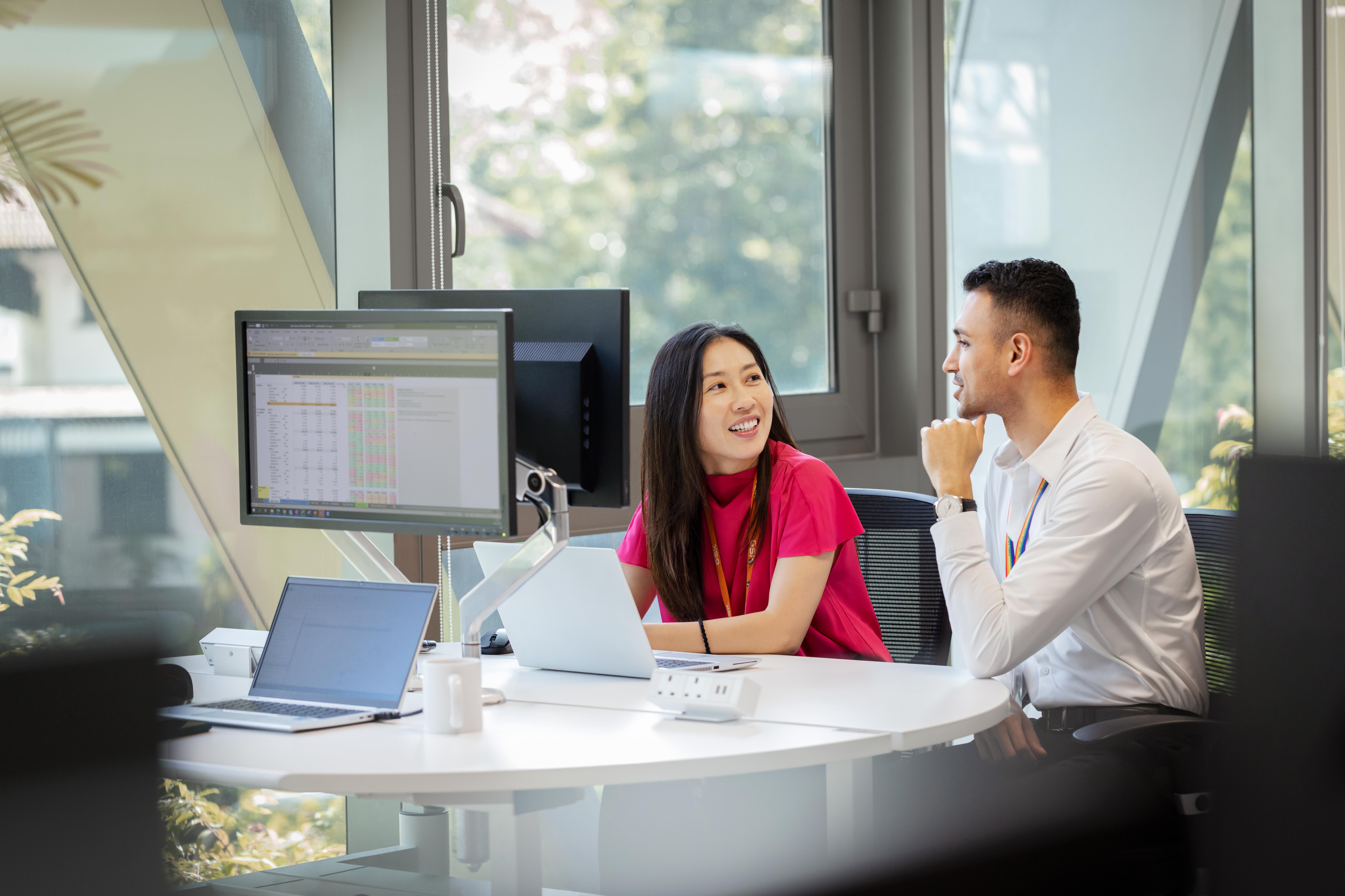 Two coworkers at a round desk discussing work while using laptops and monitors in a bright office.