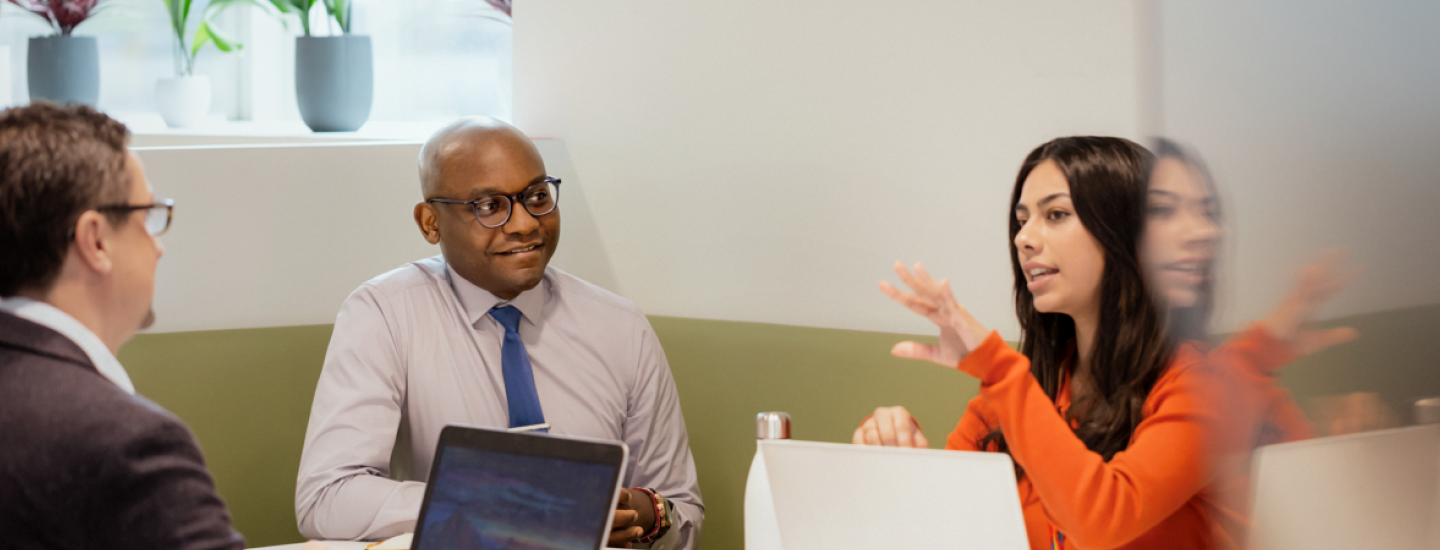 Three people around a meeting table with laptops and mugs; one person gestures while others listen, with potted plants on the windowsill behind.
