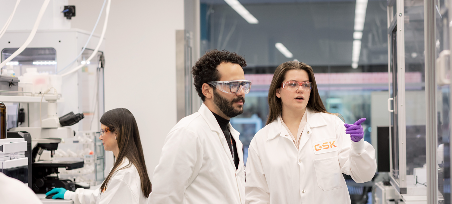 Researchers in lab coats and gloves working together in a laboratory, with one person pointing toward equipment.
