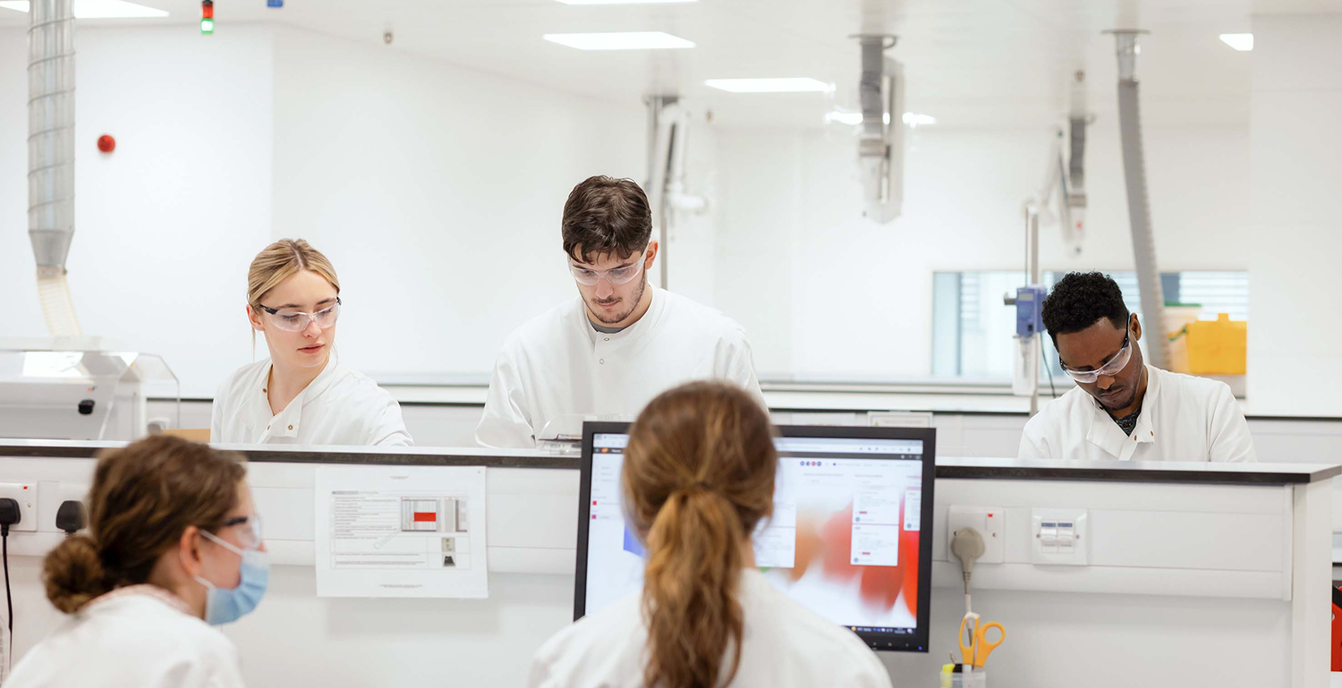 Five lab staff in white coats at benches; two at a central monitor reviewing data while three prepare equipment.