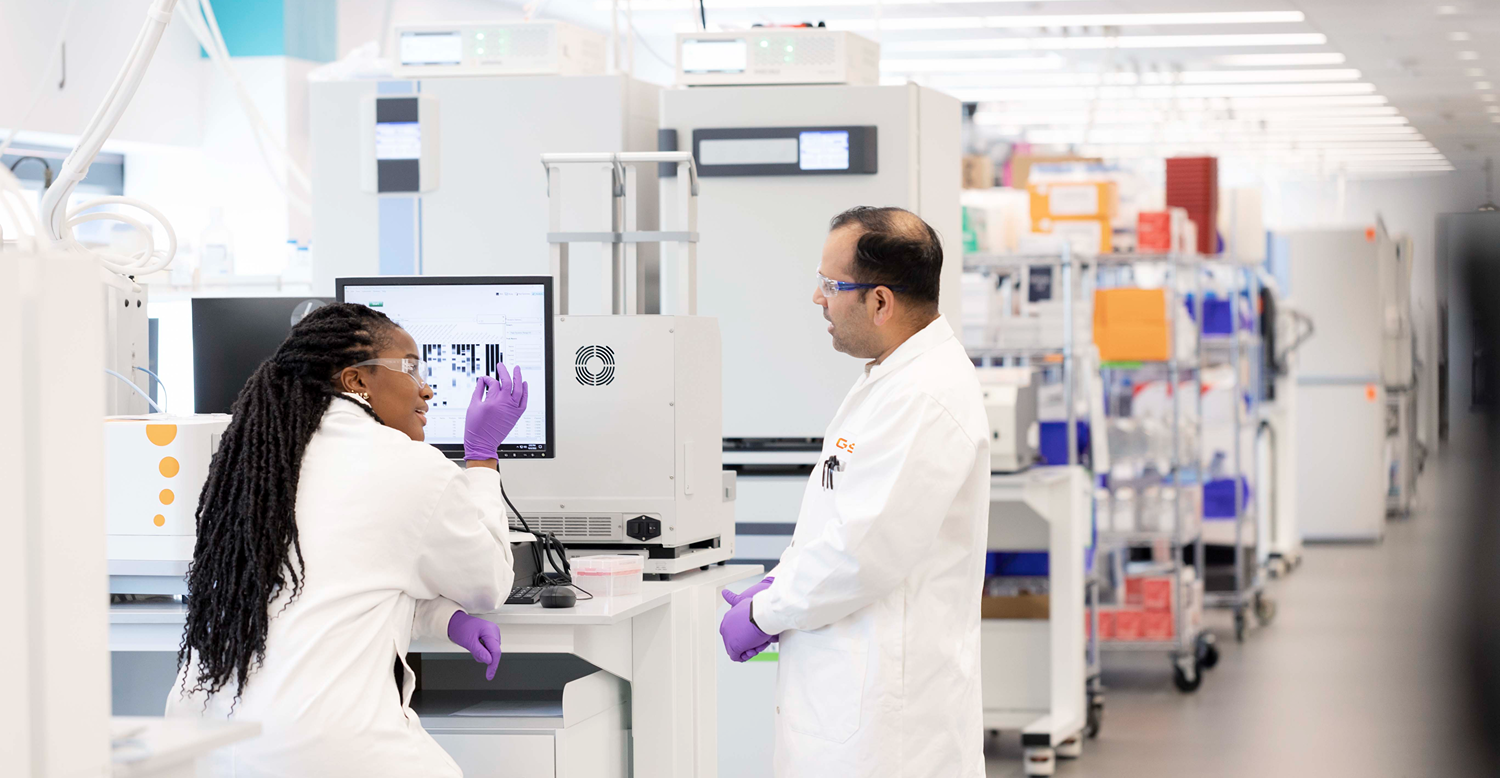 Two lab staff in white coats and purple gloves review results on a monitor while standing at a workstation in a bright lab.