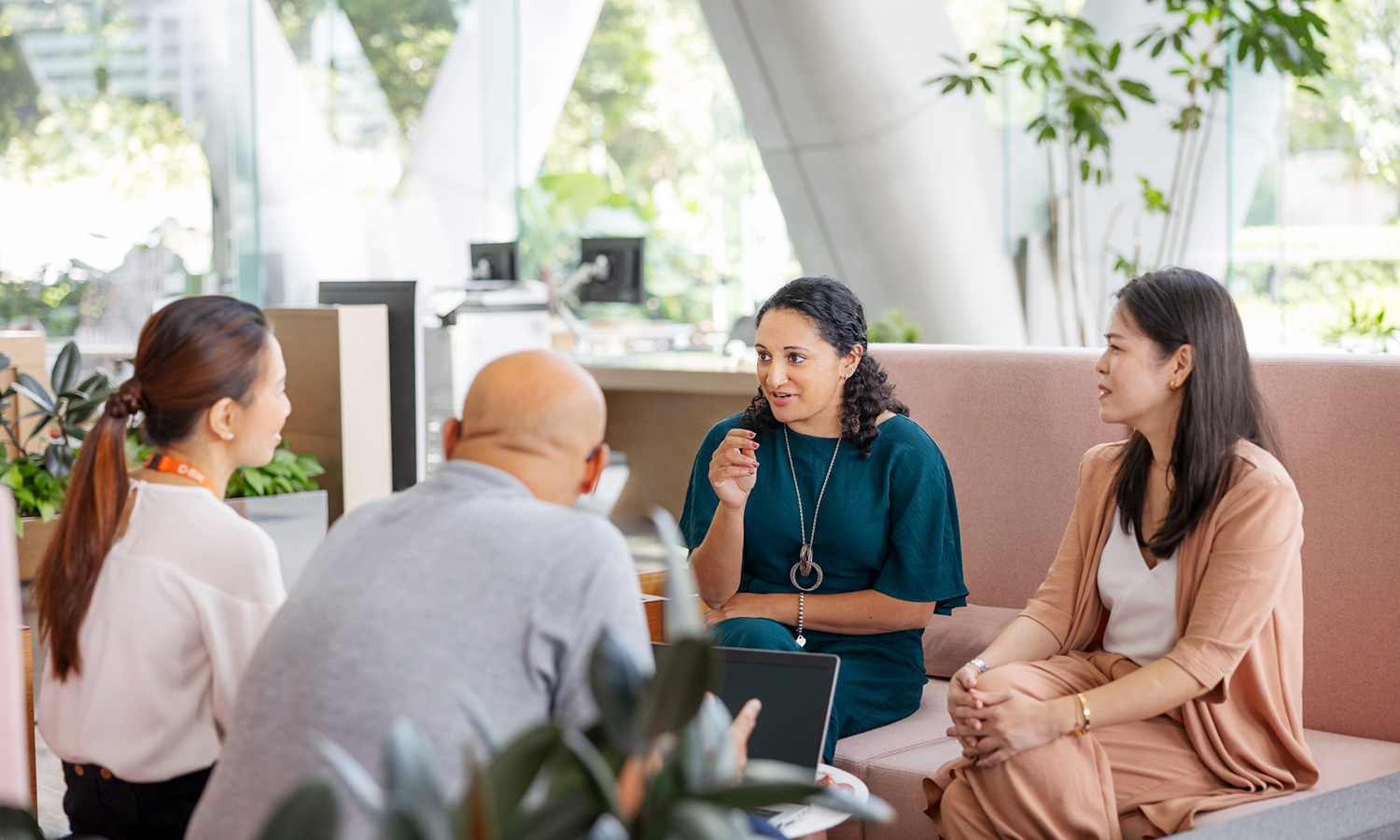  Four colleagues sit around a table in a common space, one person speaking while others listen