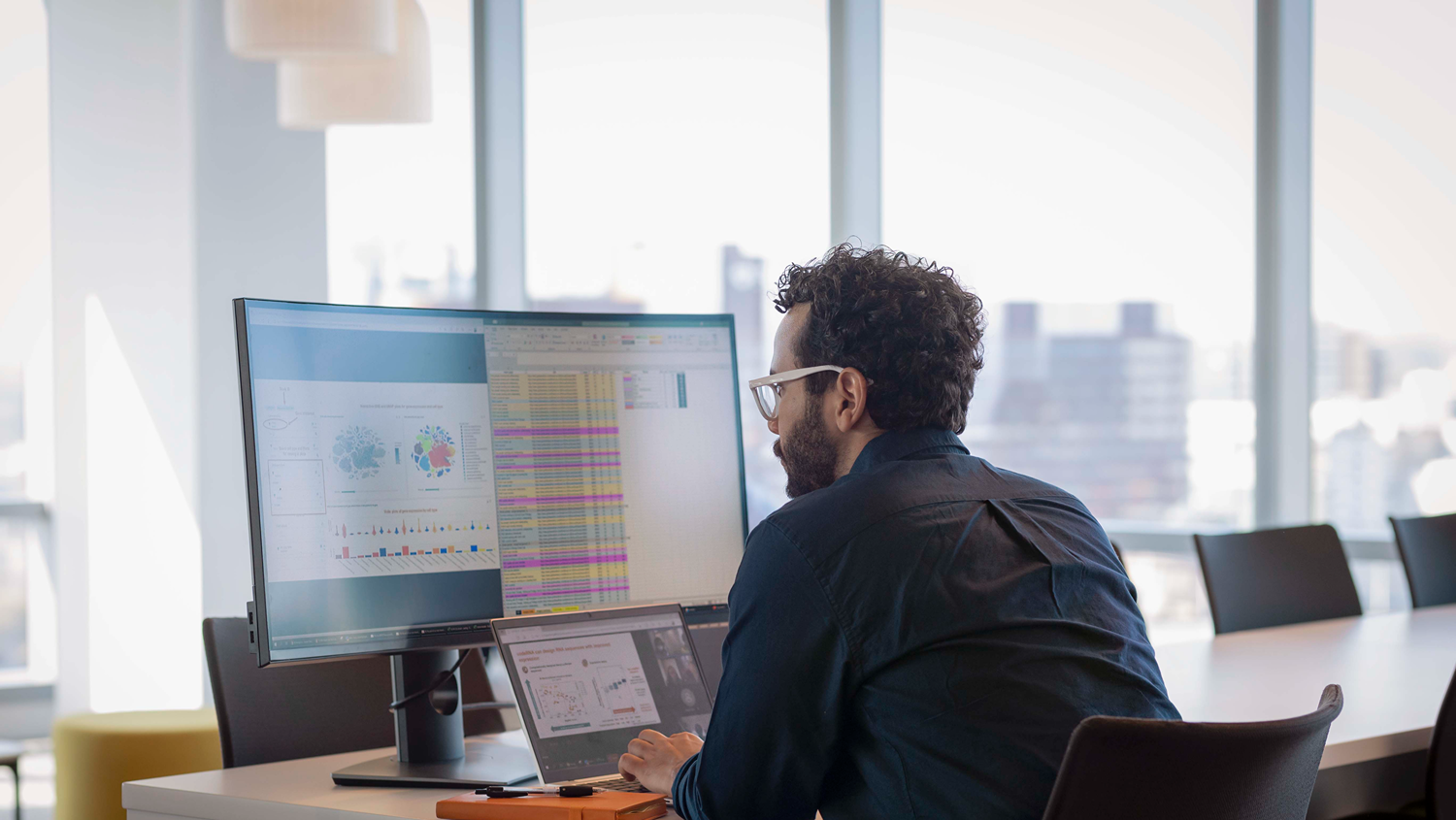 Person at a desk working on a laptop with a large monitor showing charts and a spreadsheet in a bright office.