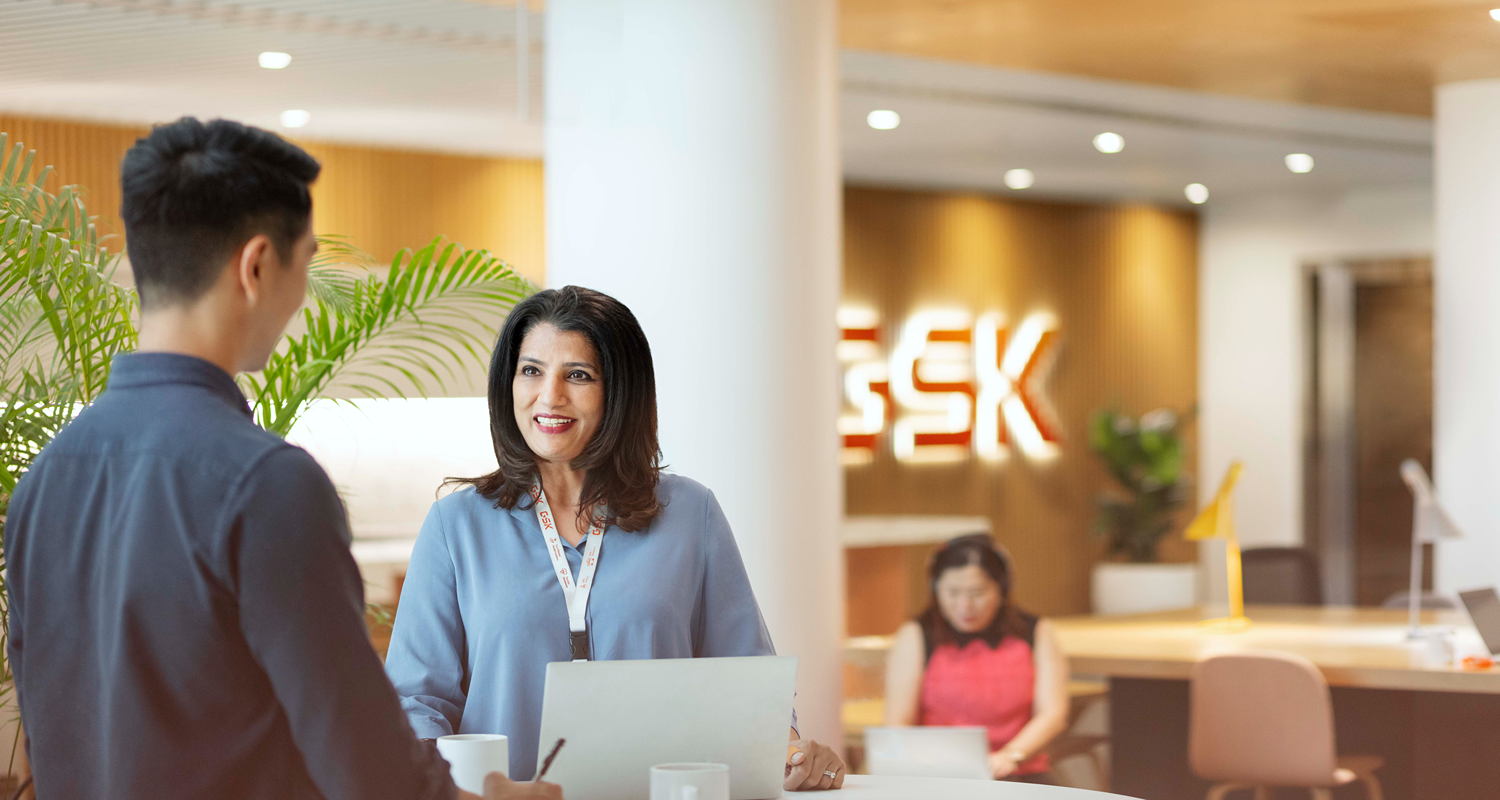 Two people talk at a round reception table in a bright area with a backlit logo on the wall and another person working in the background.