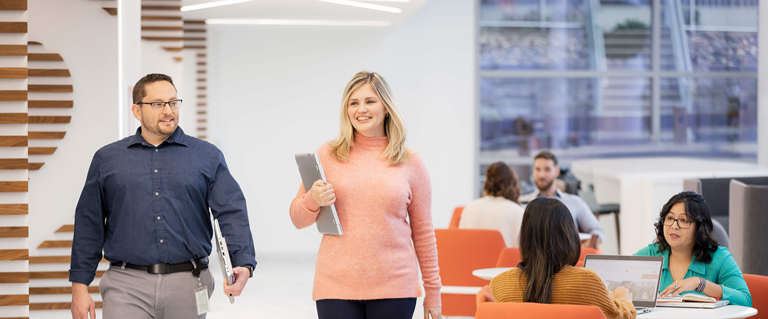 Two individuals walking in an open office space, holding laptops, while others work and collaborate at tables with orange chairs.