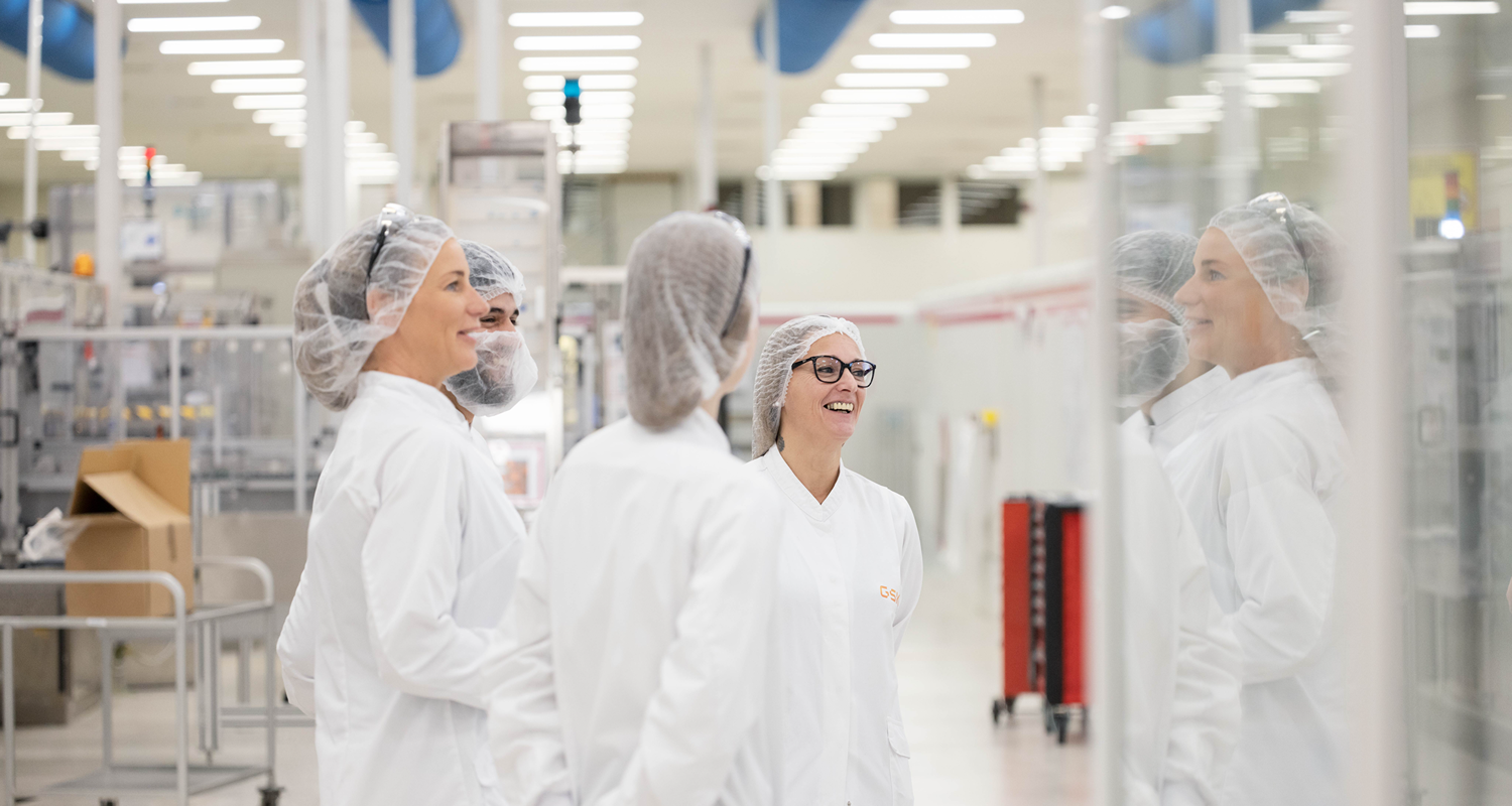Four people in lab coats and hairnets stand together in a clean manufacturing area.