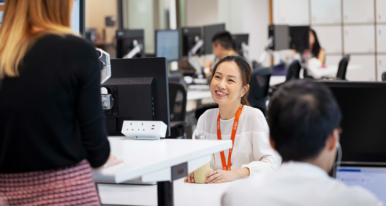 Two colleagues talk at a high desk in an open-plan office while others work at nearby monitors.