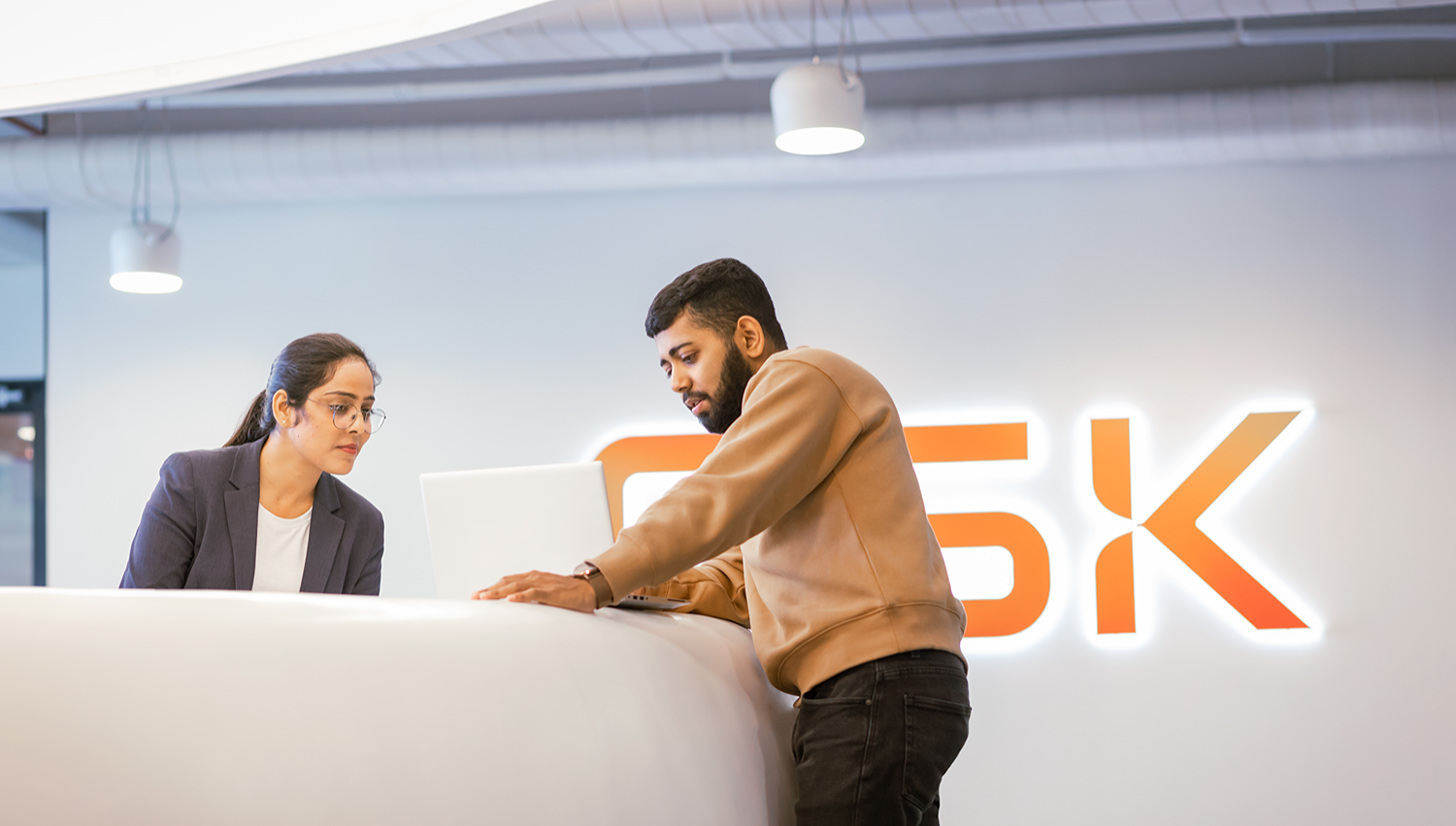 Two people interacting at a desk with a laptop. GSK logo illuminated on the wall in the background.