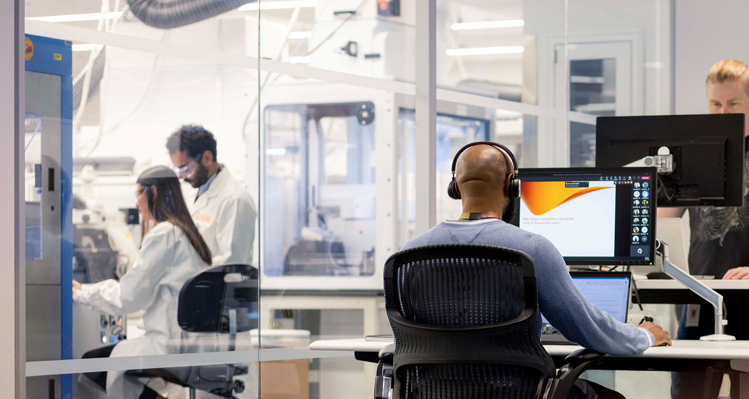 Person wearing headphones works at a desk with two monitors in a glass-walled office overlooking a lab.