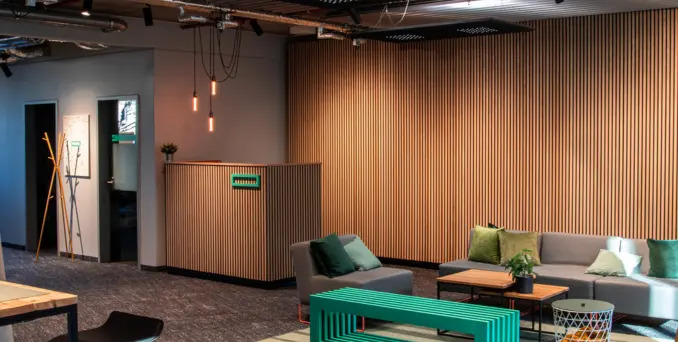 Modern office lounge with a gray sofa, green bench, and coffee table. Vertical wooden slats cover the walls. Pendant lights hang from the ceiling, and there are plants and wall art adding a cozy atmosphere.