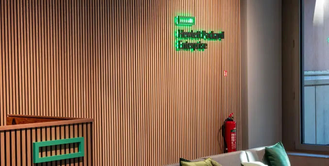 A modern office space with a wooden slat wall, green Hewlett Packard Enterprise logo, a fire extinguisher, a window to the right, and part of a white sofa with green cushions in the foreground.
