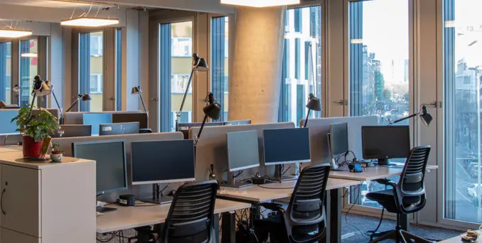 Modern open-plan office with several empty desks, black chairs, computer monitors, desk lamps, and large windows letting in natural light. A potted plant sits on a white cabinet in the foreground.