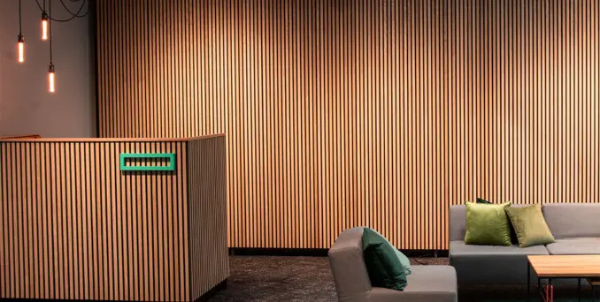 Modern office lobby with light wood slat walls, grey sofas, and green accent pillows. Hanging pendant lights are visible, creating a warm and minimalist atmosphere.