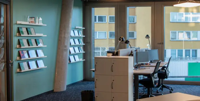 Modern office with desks, chairs, lamps, and computer monitors near large windows. Shelves with books line a green wall. Outside, yellow apartment buildings are visible. The space is tidy and well-lit with natural light.
