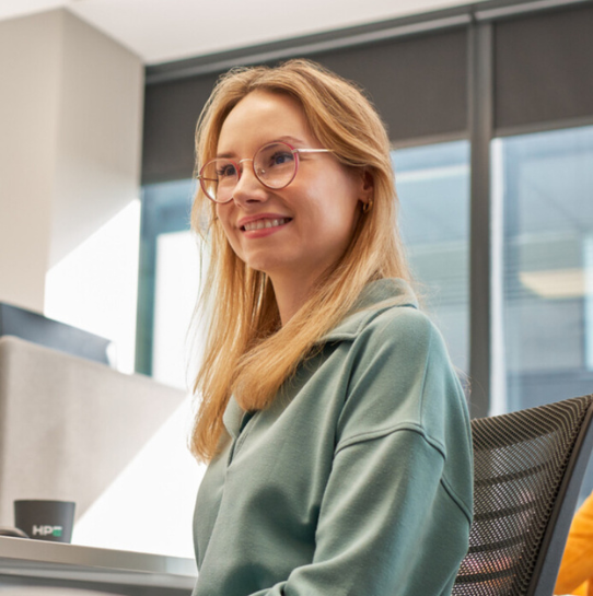 A young woman with long blonde hair, wearing glasses and a teal sweater, sits on an office chair and smiles. Large windows and office furniture are visible in the background.