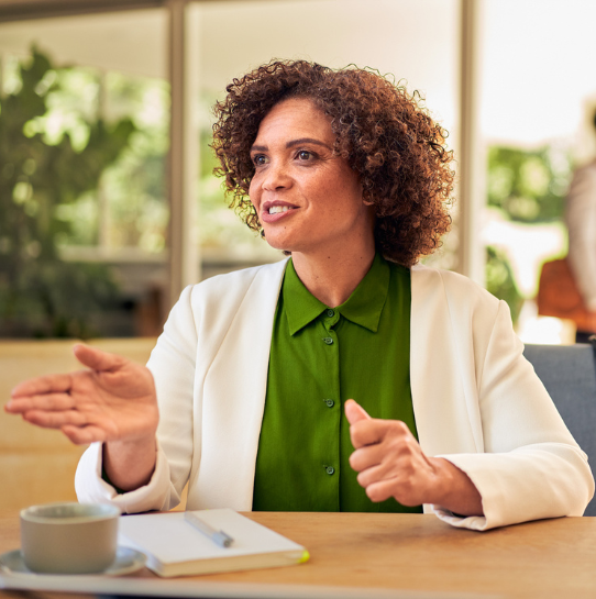 A woman with curly hair, wearing a white blazer and green shirt, sits at a table with a notebook and coffee cup, gesturing and smiling in a bright indoor setting with plants in the background.