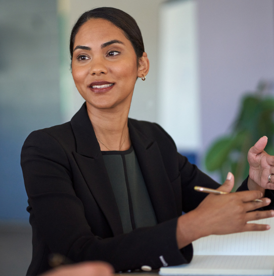 A woman in a black blazer sits at a table, holding a pen and gesturing with her hand, appearing engaged in conversation. A notebook is in front of her, and a plant is visible in the background.