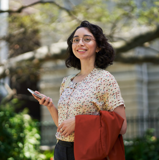 A woman with curly dark hair and glasses smiles while holding a smartphone and an orange blazer outdoors. She wears a patterned blouse and stands in front of greenery and a blurred building.