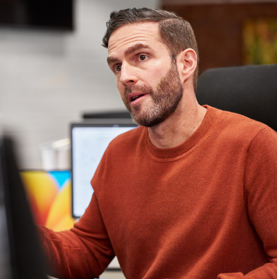 A man with short dark hair and a beard, wearing an orange sweater, sits at a desk in an office setting, speaking and gesturing with his hand. Computer monitors are visible in the background.