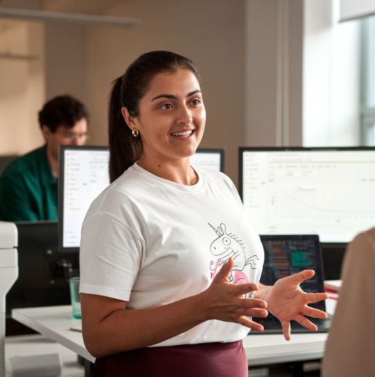 A woman wearing a white unicorn t-shirt is speaking and gesturing with her hands in an office with computer monitors and another person working in the background.