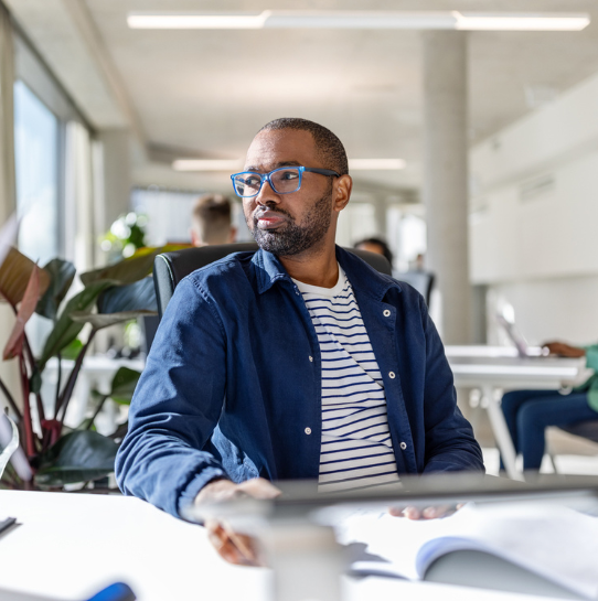 A man with glasses and a beard, wearing a blue jacket over a striped shirt, sits at a bright office desk looking thoughtfully to the side. Plants and other people working are visible in the background.