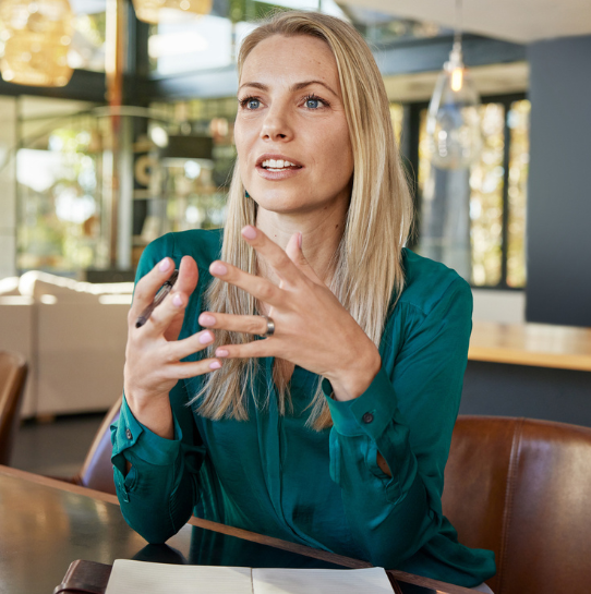 A woman with long blonde hair in a green blouse sits at a table, gesturing with her hands as she speaks. An open notebook and pen are in front of her in a modern, well-lit office space.