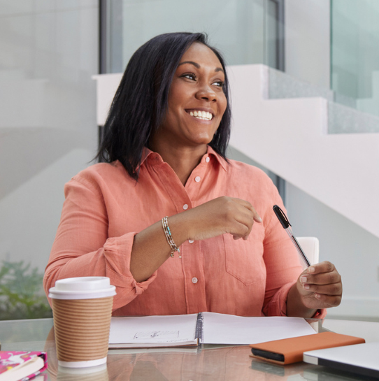 A woman in a coral shirt sits at a desk, smiling and holding a pen. A notebook, a paper coffee cup, and a closed tablet are on the desk in front of her. A modern staircase is in the background.