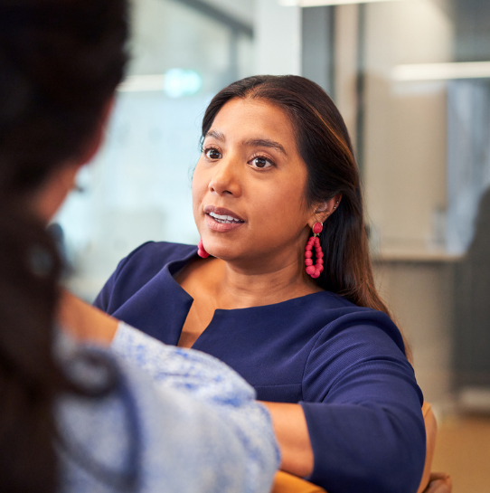 A woman with long dark hair and pink earrings, wearing a blue top, sits and listens attentively to another person in an office setting. The second person is out of focus in the foreground.