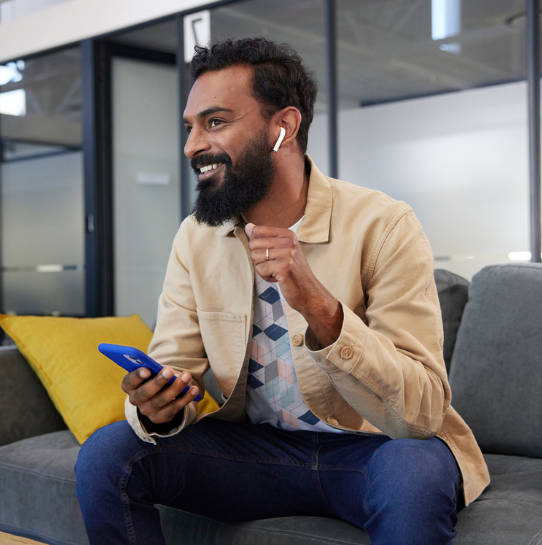 A man with a beard, wearing a tan jacket and wireless earbuds, sits on a couch smiling while holding a blue smartphone. He appears to be in a modern office or lounge area.