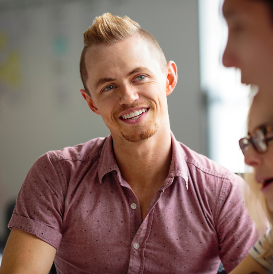 A smiling man with short blond hair and a trimmed beard wears a light pink short-sleeve button-up shirt, sitting indoors and engaging in conversation with two other people, who are partially visible.