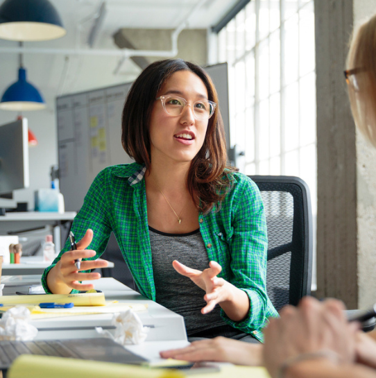 A woman wearing glasses and a green plaid shirt gestures while talking to a colleague at a modern office desk, with papers, pens, and crumpled notes in front of her and large windows in the background.