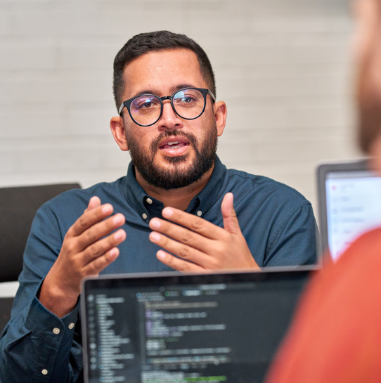 A man wearing glasses and a dark shirt is speaking and gesturing with his hands during a discussion. He is sitting at a table with laptops open, and code is visible on a laptop screen in the foreground.