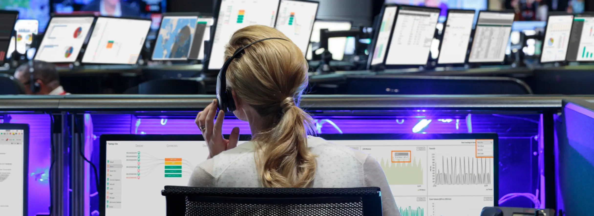 A woman with a headset sits at a workstation with multiple monitors displaying charts and data in a modern control room or operations center.