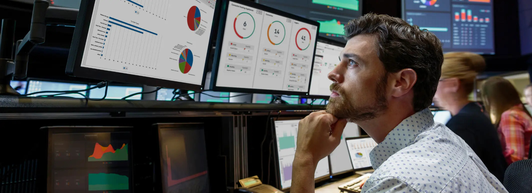 A man with a beard looks thoughtfully at several computer monitors displaying charts, graphs, and data visualizations in a modern office setting.