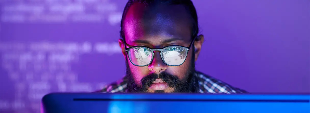 A person with glasses and a beard sits in front of a computer screen, with code reflected in their lenses and a purple background visible behind them.
