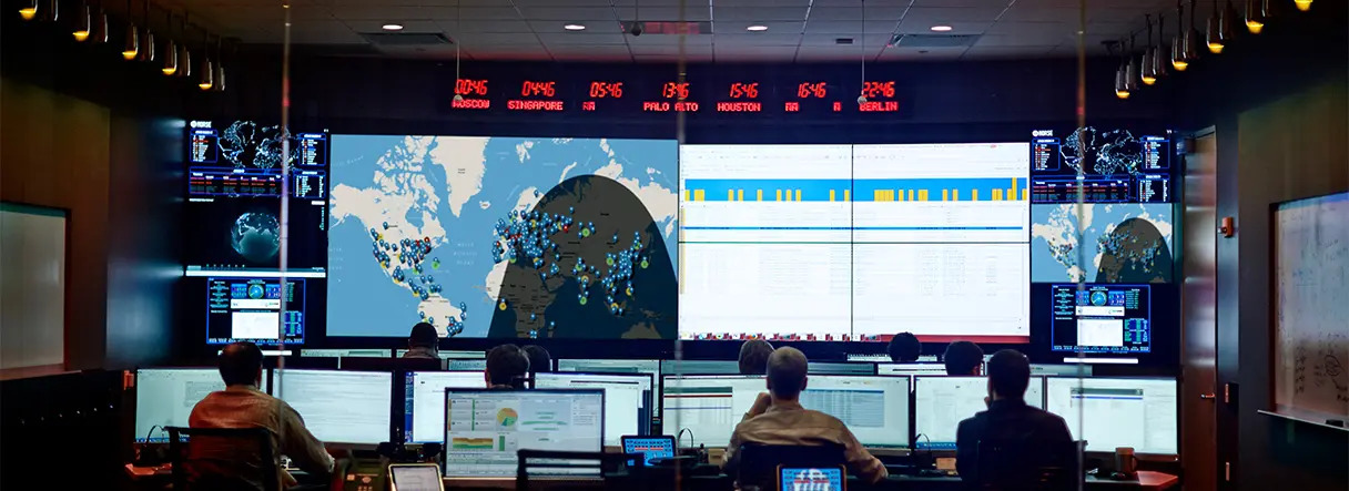 People sit at computer workstations in a dark control room, facing large screens displaying a world map, data charts, and cybersecurity information. Red digital clocks show world times above the main screens.