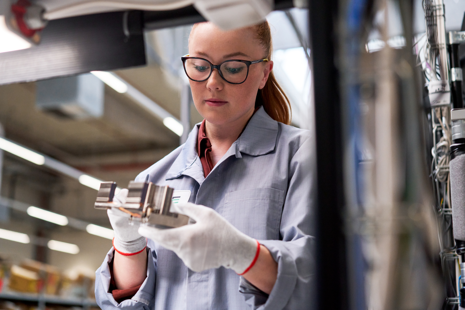 A woman wearing glasses, a lab coat, and white gloves inspects a metal component in an industrial or laboratory setting with shelves and equipment in the background.