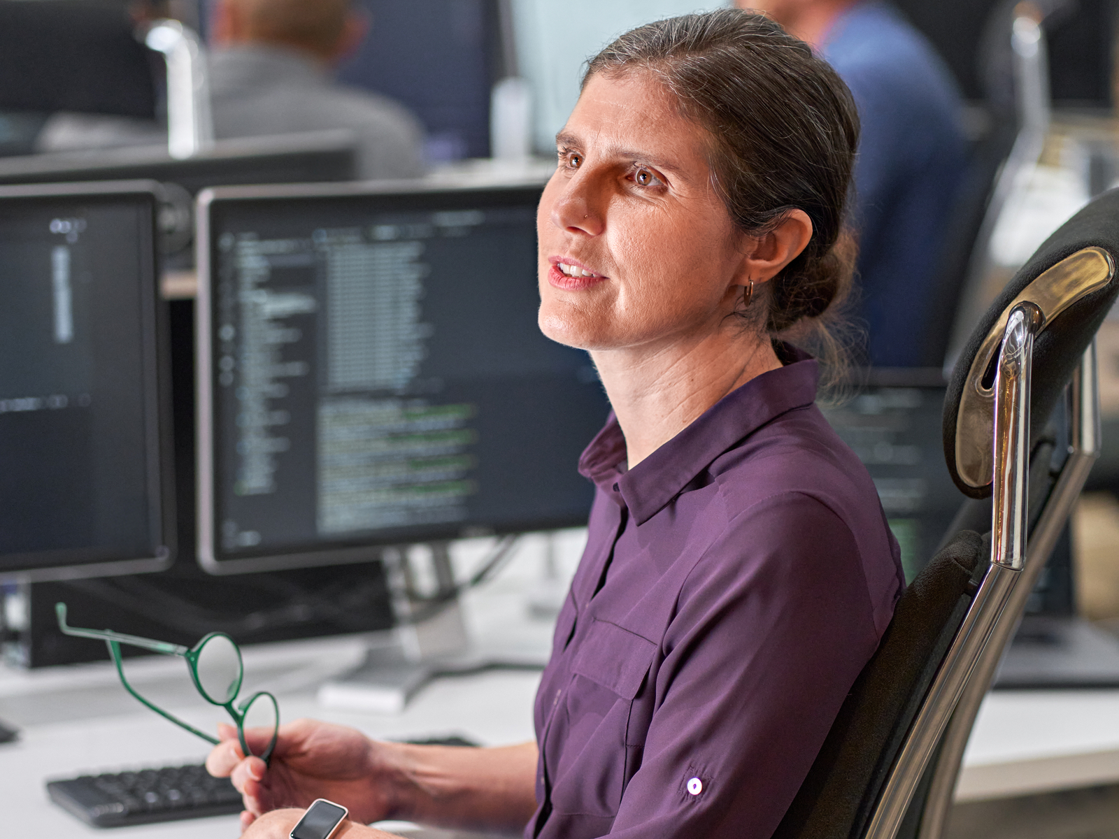 A woman with brown hair tied back, wearing a purple shirt, sits at a desk holding glasses and a phone. Computer monitors with code are visible in the background of a modern office space.