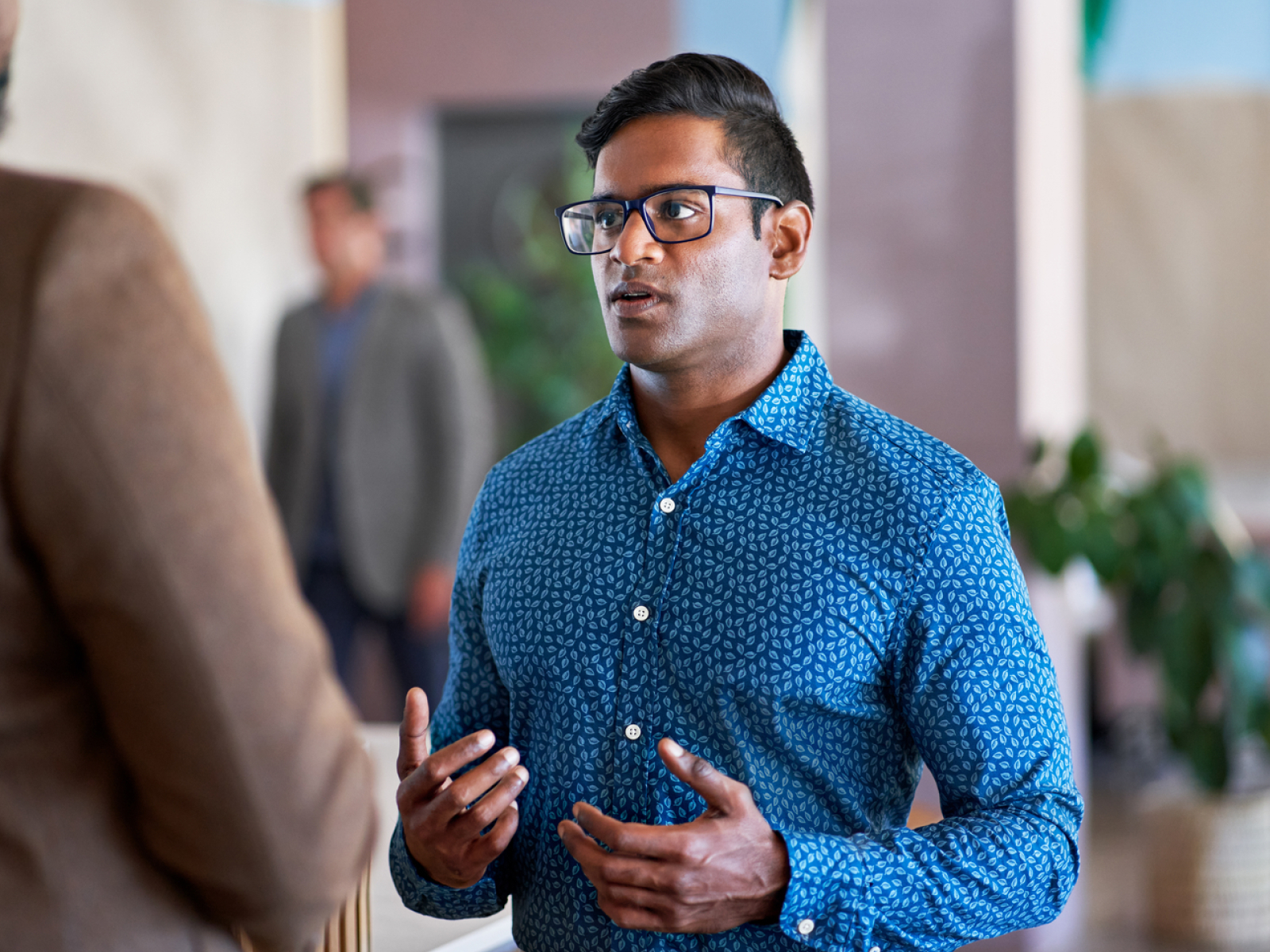 A man wearing glasses and a blue patterned shirt is speaking, gesturing with his hands. He is indoors with a blurred background of people and plants.