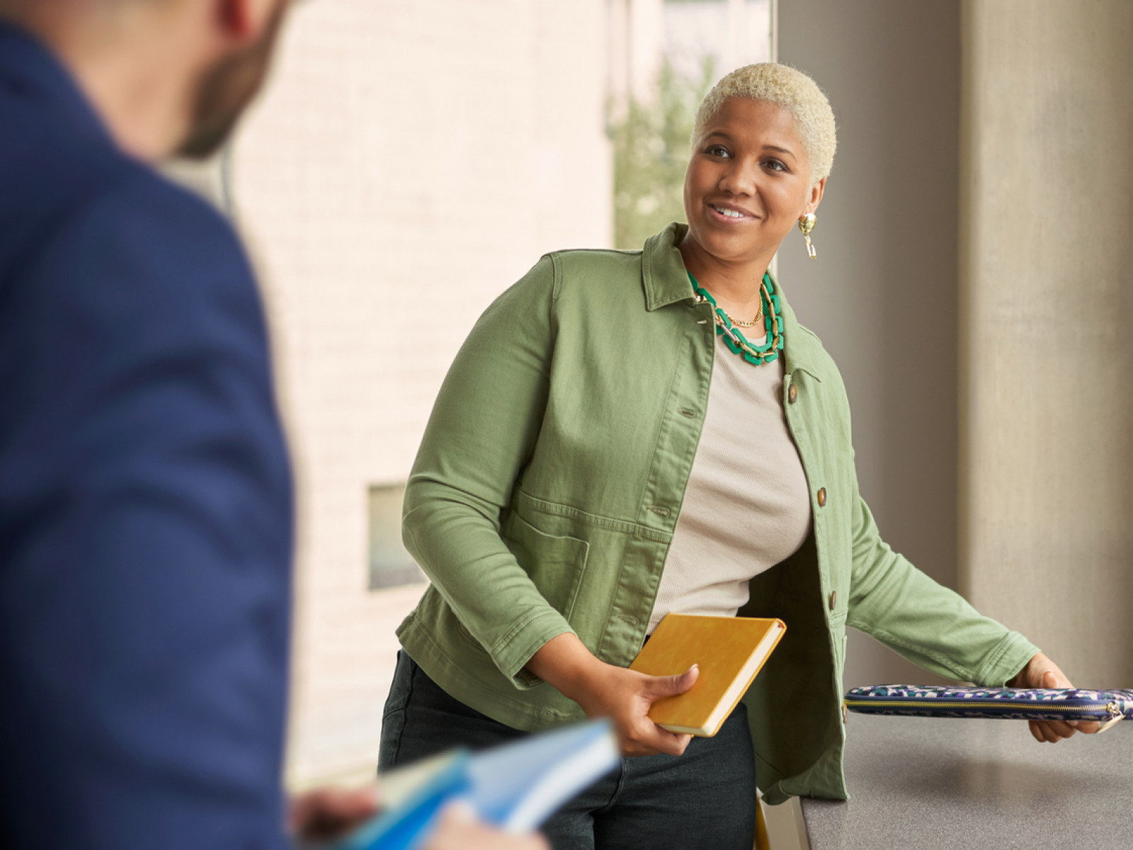 A woman with short blonde hair, wearing a green jacket and beige shirt, smiles and holds a yellow notebook and a laptop while standing by a window, talking to a man in a blue jacket.